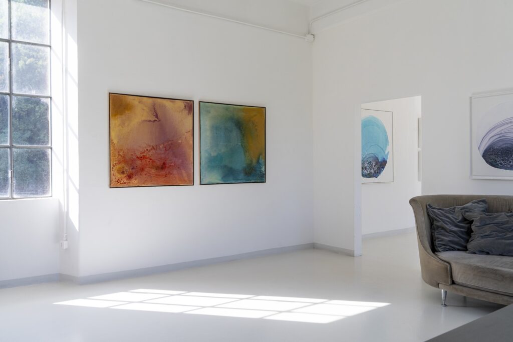 Sala della galleria con due dipinti di Franza, luce naturale dalla finestra e divano grigio in velluto